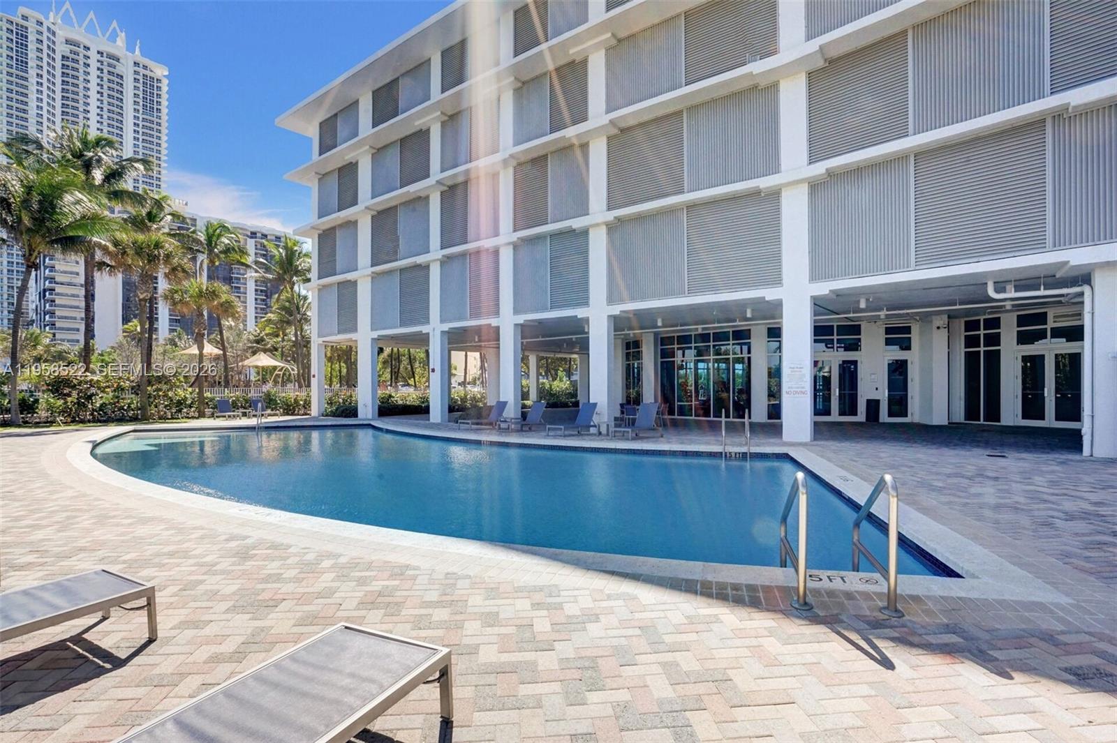 An outdoor swimming pool sits in front of a multi-story building with a paved deck — 6515 Collins Ave # 901, Miami Beach FL 33141, MIAMI BEACH, FL