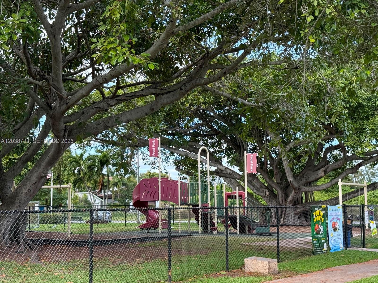 A fenced playground with a red slide sits under large trees at Frost Park — 610 NE 2nd St # 305, Dania Beach FL 33004, DANIA BEACH, FL