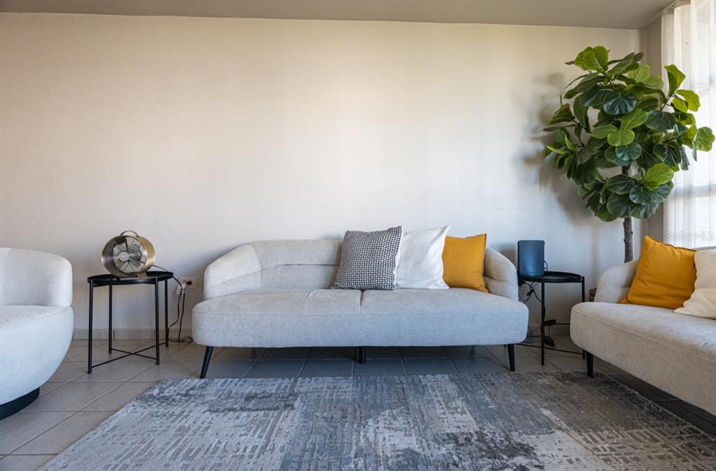 A living room features a light gray sofa, two accent chairs, and a large potted plant — Apto 14-402 COSTA BRAVA, CEIBA, PR
