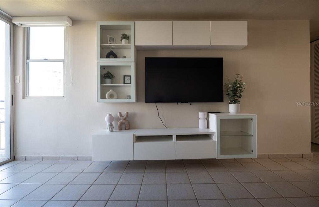 A living room features a wall-mounted television, white cabinetry, and light-colored tiled flooring — Apto 14-402 COSTA BRAVA, CEIBA, PR