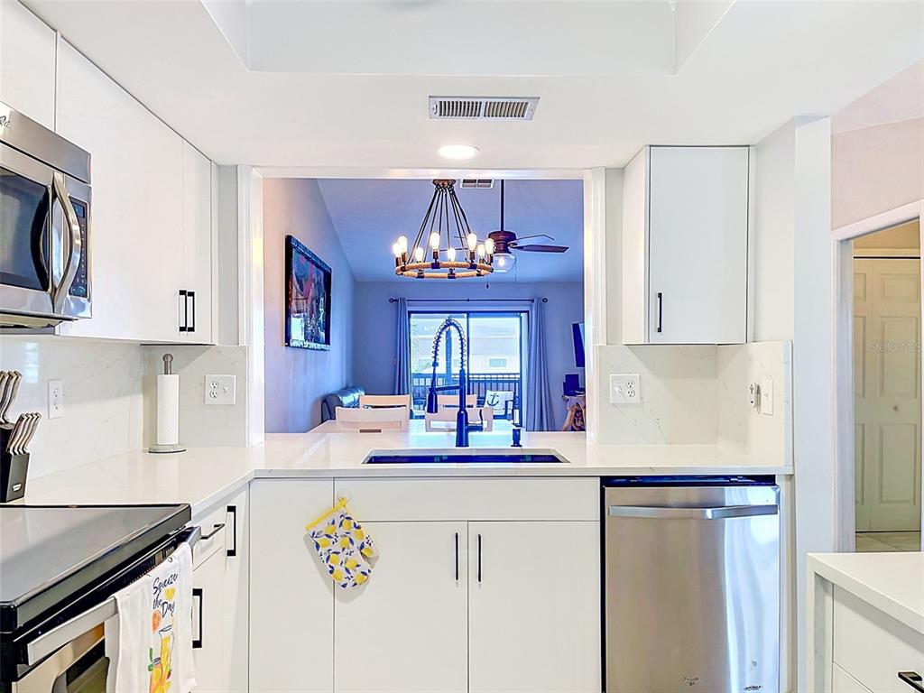 A white kitchen features stainless steel appliances, quartz countertops, and a pass-through window to the dining area — 460 S PAULA DR UNIT 107, DUNEDIN, FL