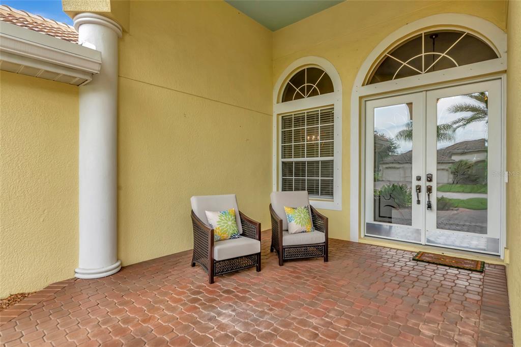 A covered porch with brick pavers, two wicker chairs, and double glass doors — 14711 2ND AVENUE CIRCLE NE, BRADENTON, FL
