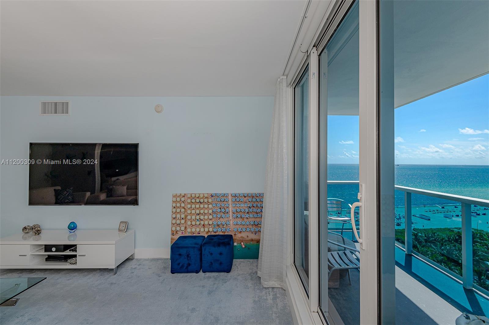 A living room features a wall-mounted television, blue ottomans, and a balcony overlooking the ocean — 2301 Collins Ave # 1506, Miami Beach FL 33139, MIAMI BEACH, FL