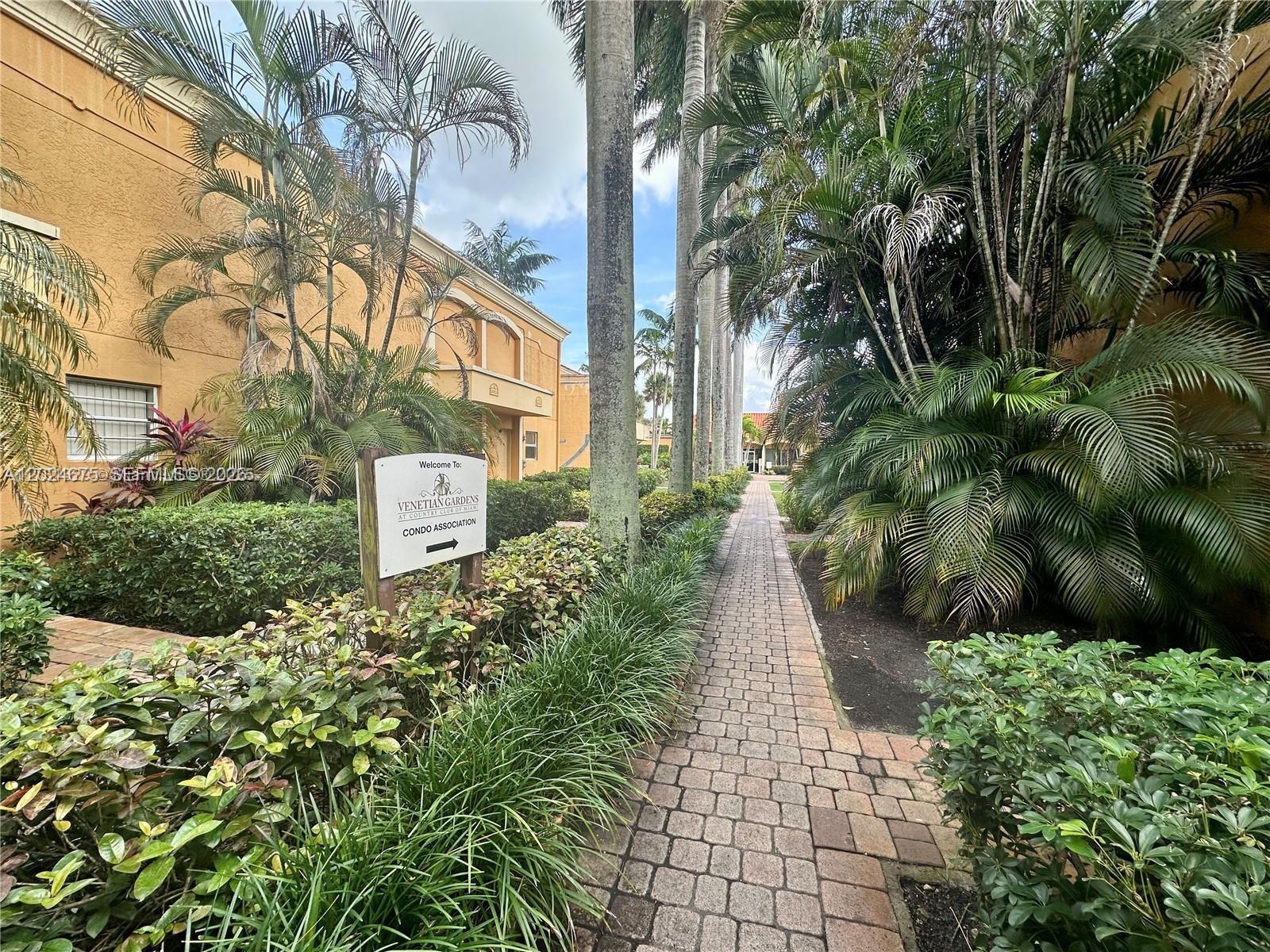 A brick walkway leads through a landscaped courtyard between two yellow residential buildings — 18111 NW 68th Ave # F203, Hialeah FL 33015, HIALEAH, FL