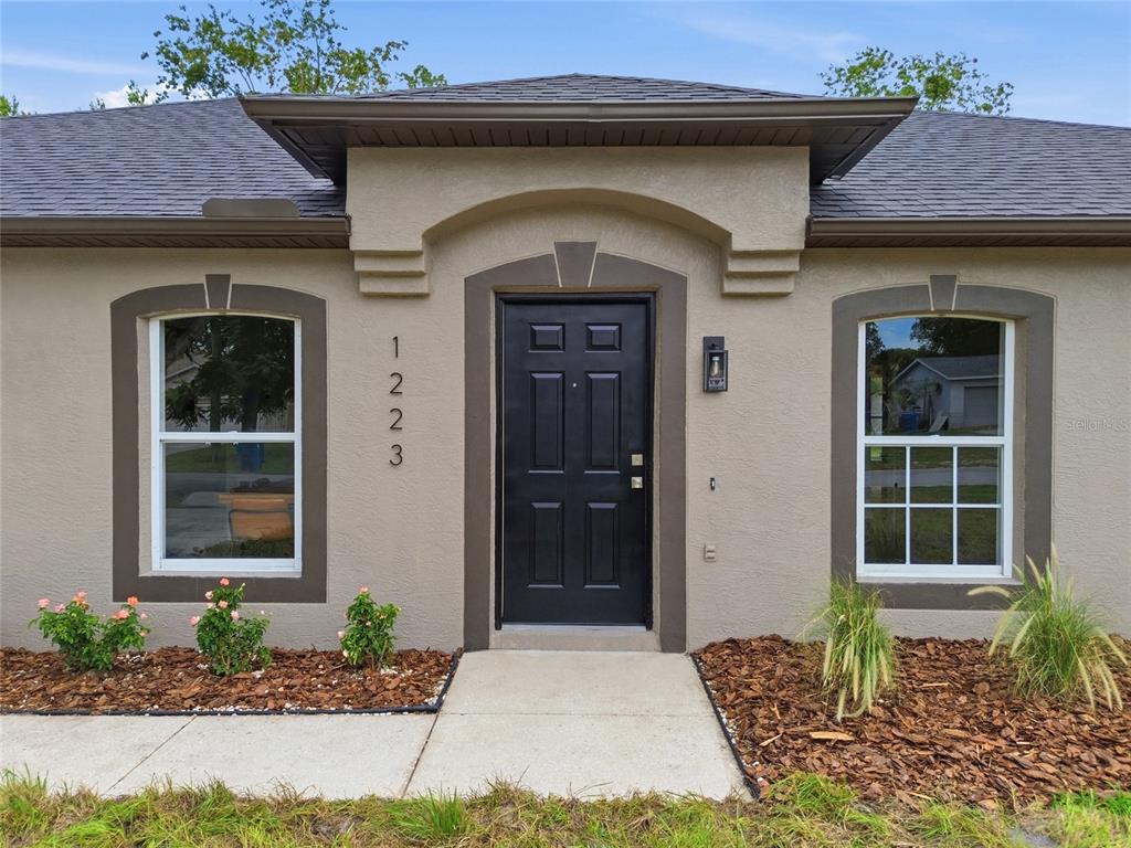 The front exterior of a house features a black door, two windows, and landscaping — 1223 GATEWOOD AVENUE, SPRING HILL, FL