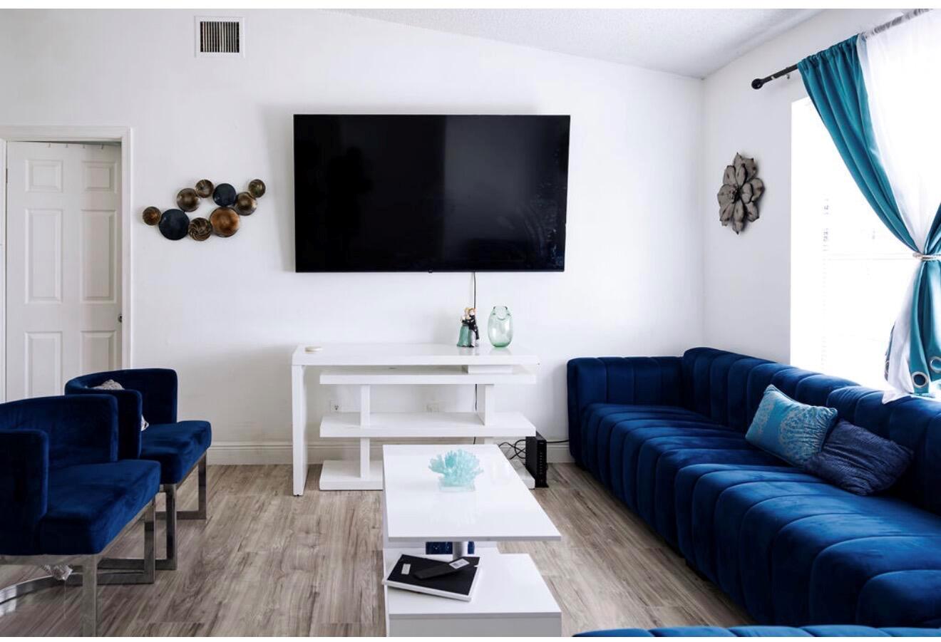A living room features a wall-mounted television, a white console table, and blue velvet seating — 5437 Greenwood Drive, Delray Beach FL 33484, DELRAY BEACH, FL