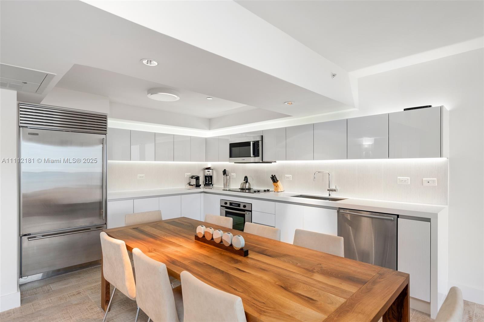 A modern kitchen features white cabinetry, stainless steel appliances, and a large wooden dining table — 102 24th St # 1219, Miami Beach FL 33139, MIAMI BEACH, FL