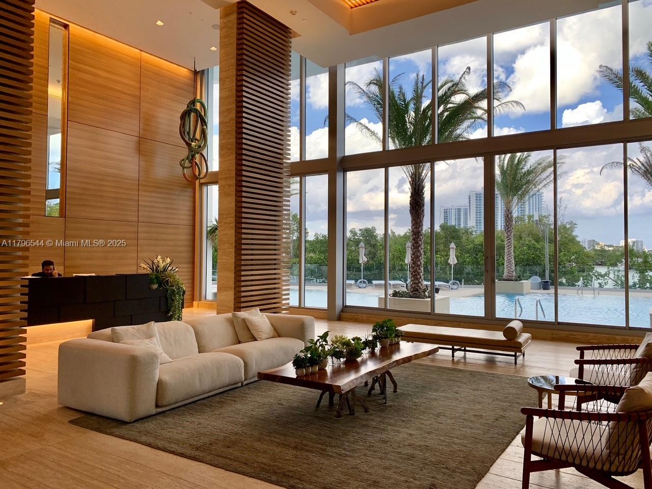 A modern lobby features a large sofa, wood-slat columns, and floor-to-ceiling windows overlooking a pool — 16385 Biscayne Blvd, North Miami Beach FL 33160, NORTH MIAMI BEACH, FL