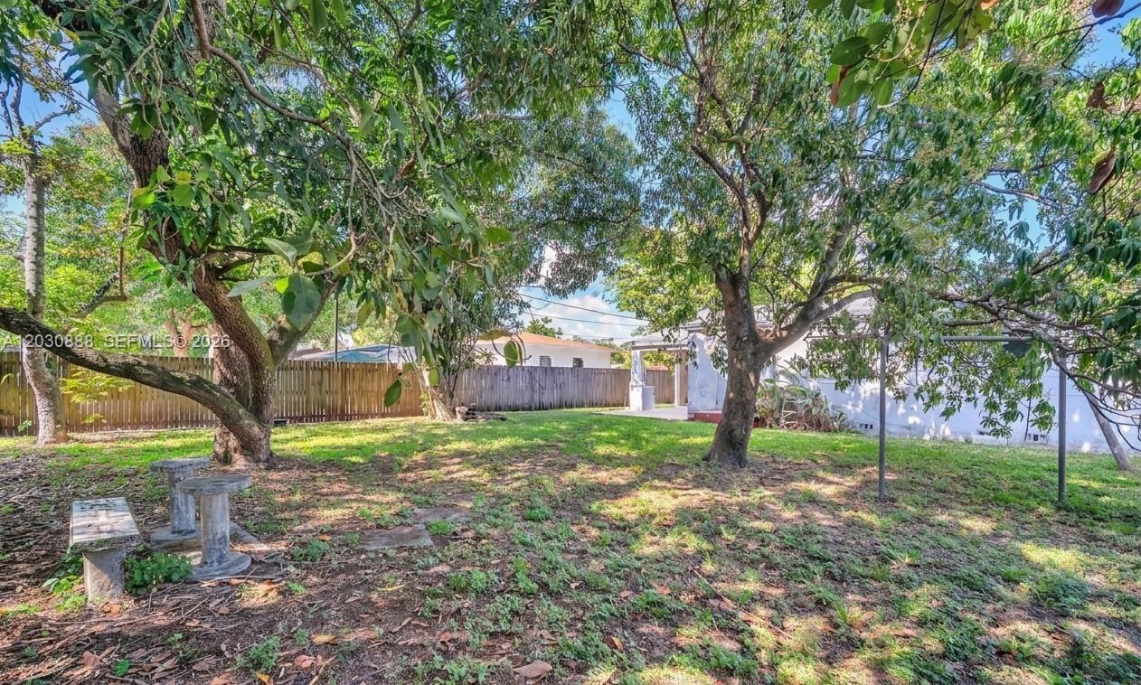 A grassy backyard features mature trees, a wooden fence, and a stone bench — 1236 Jann Ave, Opa-Locka FL 33054, OPA-LOCKA, FL