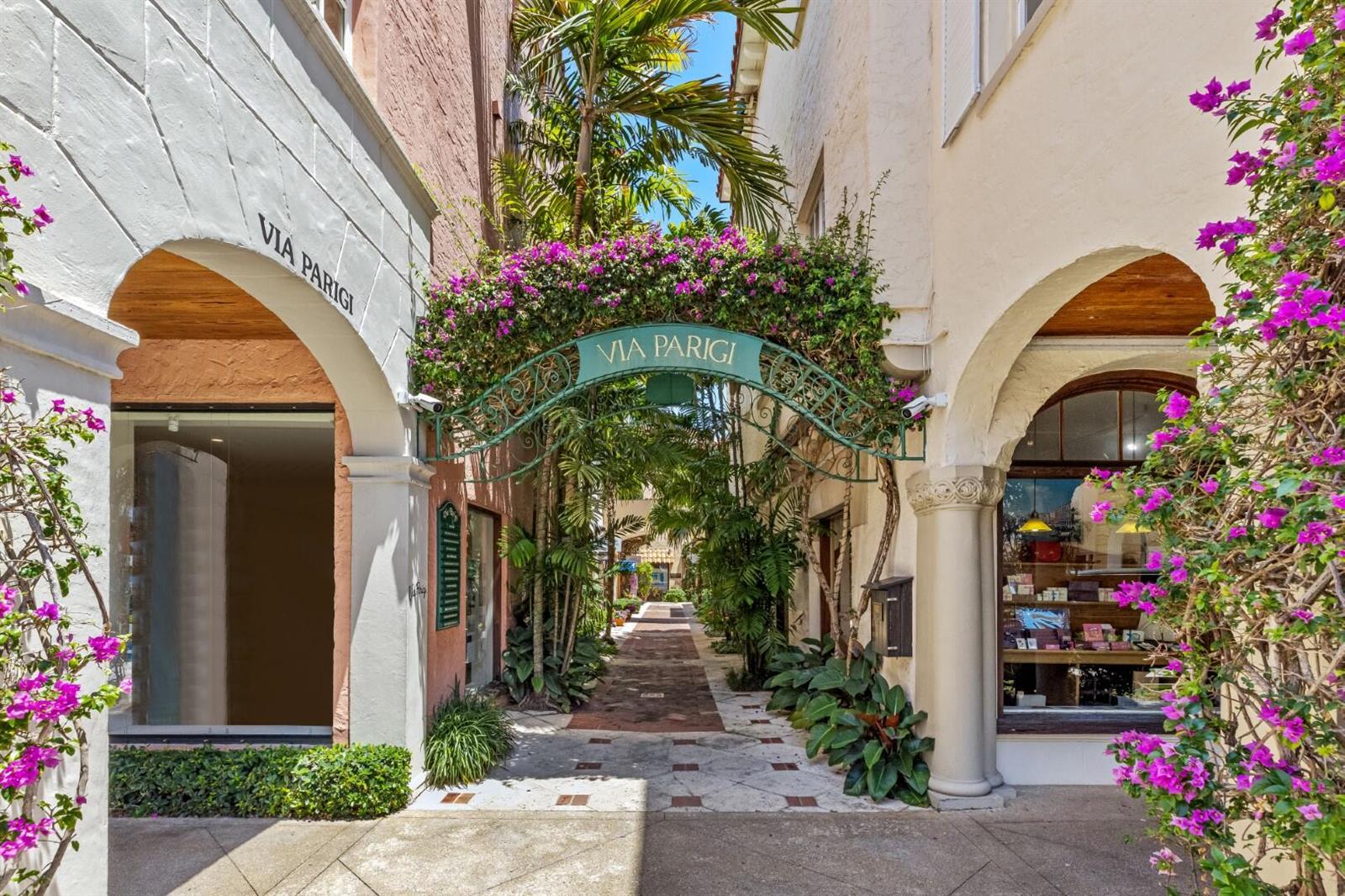 A narrow, sunlit pedestrian walkway features arched entrances, lush greenery, and a decorative metal archway — 9 Via Parigi, Palm Beach FL 33480, PALM BEACH, FL