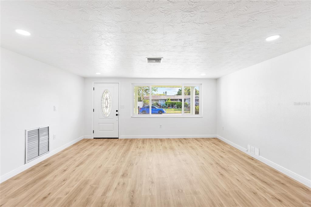 A bright, empty living room features white walls, light wood-look flooring, and a front door — 6239 7TH AVENUE, NEW PORT RICHEY, FL