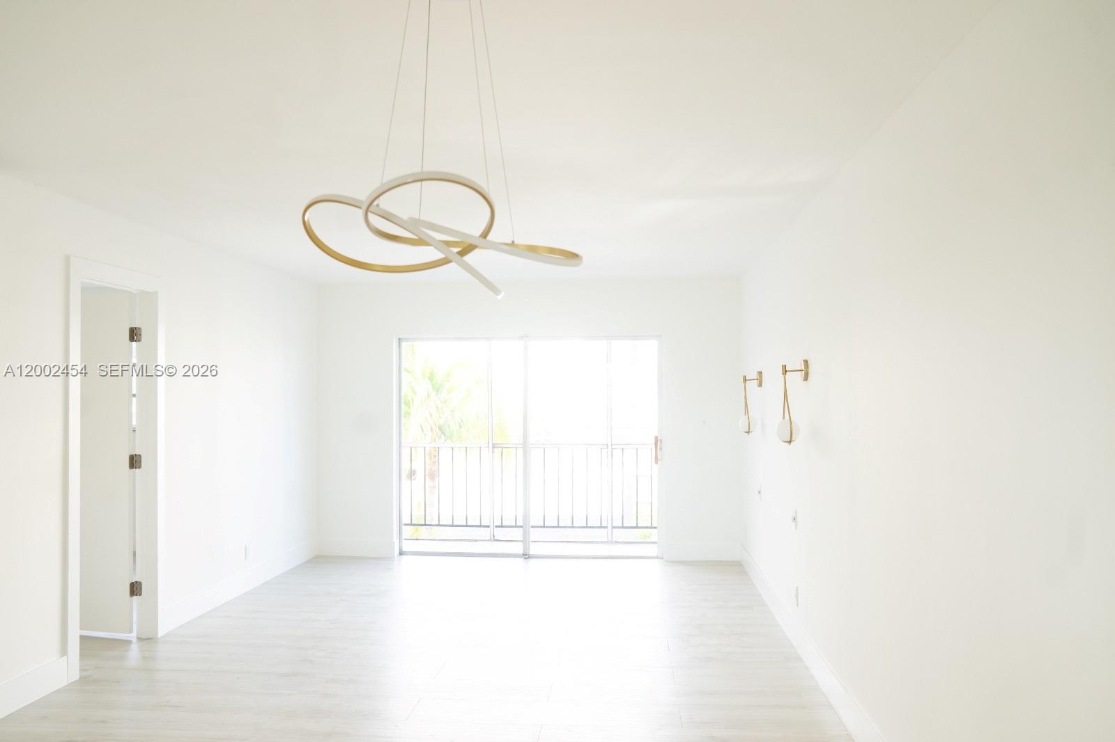 A bright, minimalist room features light wood flooring, a modern gold chandelier, and sliding doors — 220 SW 9th Ave # 306, Hallandale Beach FL 33009, HALLANDALE BEACH, FL