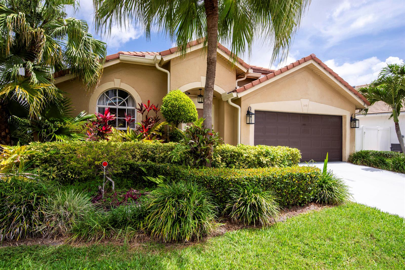 A tan, single-story house with a brown garage door and lush landscaping in the front — 149 Cypress Cove, Jupiter FL 33458, JUPITER, FL