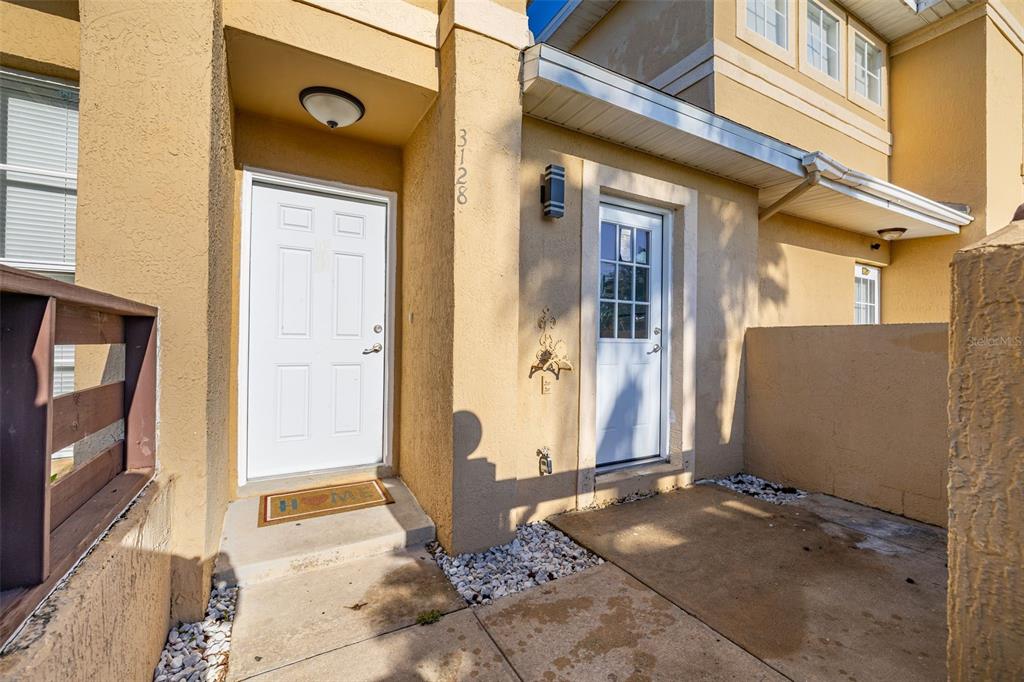 The exterior of a tan stucco home features two white doors and a concrete patio — 3128 SANDY SHORE LANE, KISSIMMEE, FL