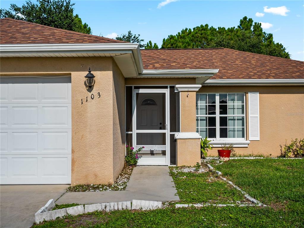 The exterior of a tan house features a white garage door, a screen door, and windows — 1103 LEE AVENUE, LEHIGH ACRES, FL