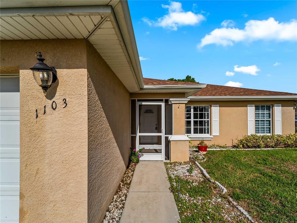 The exterior of a tan house features a concrete walkway leading to a screened front door — 1103 LEE AVENUE, LEHIGH ACRES, FL