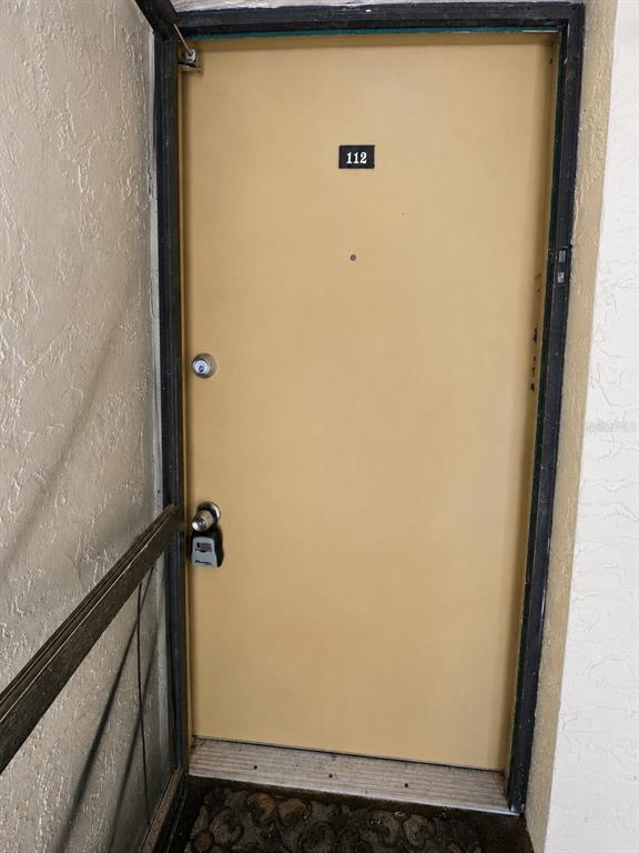 A tan apartment door labeled 112 features a lockbox on the handle and a screen door — 112 CYPRESS COURT UNIT 112, OLDSMAR, FL