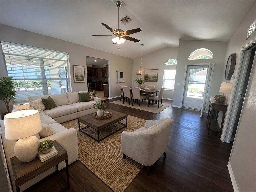 An open-concept living and dining area features vaulted ceilings, dark wood flooring, and neutral furniture — 10972 SW 69TH CIRCLE, OCALA, FL