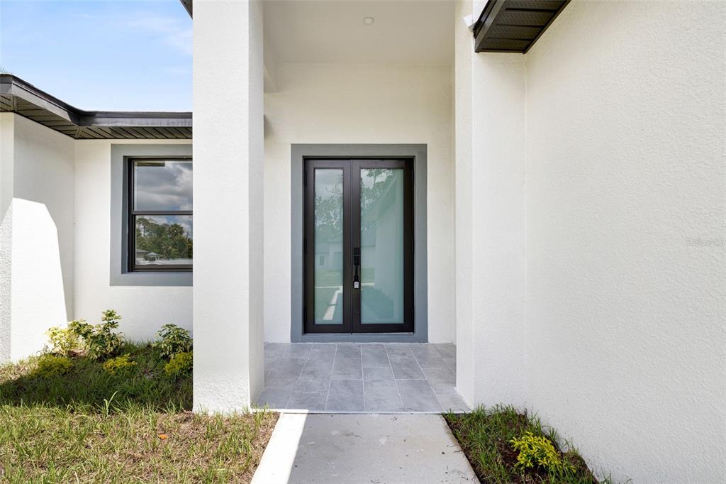 A modern home entrance features a double glass door, white stucco walls, and tiled flooring — 16290 BANYAN AVENUE, PORT CHARLOTTE, FL