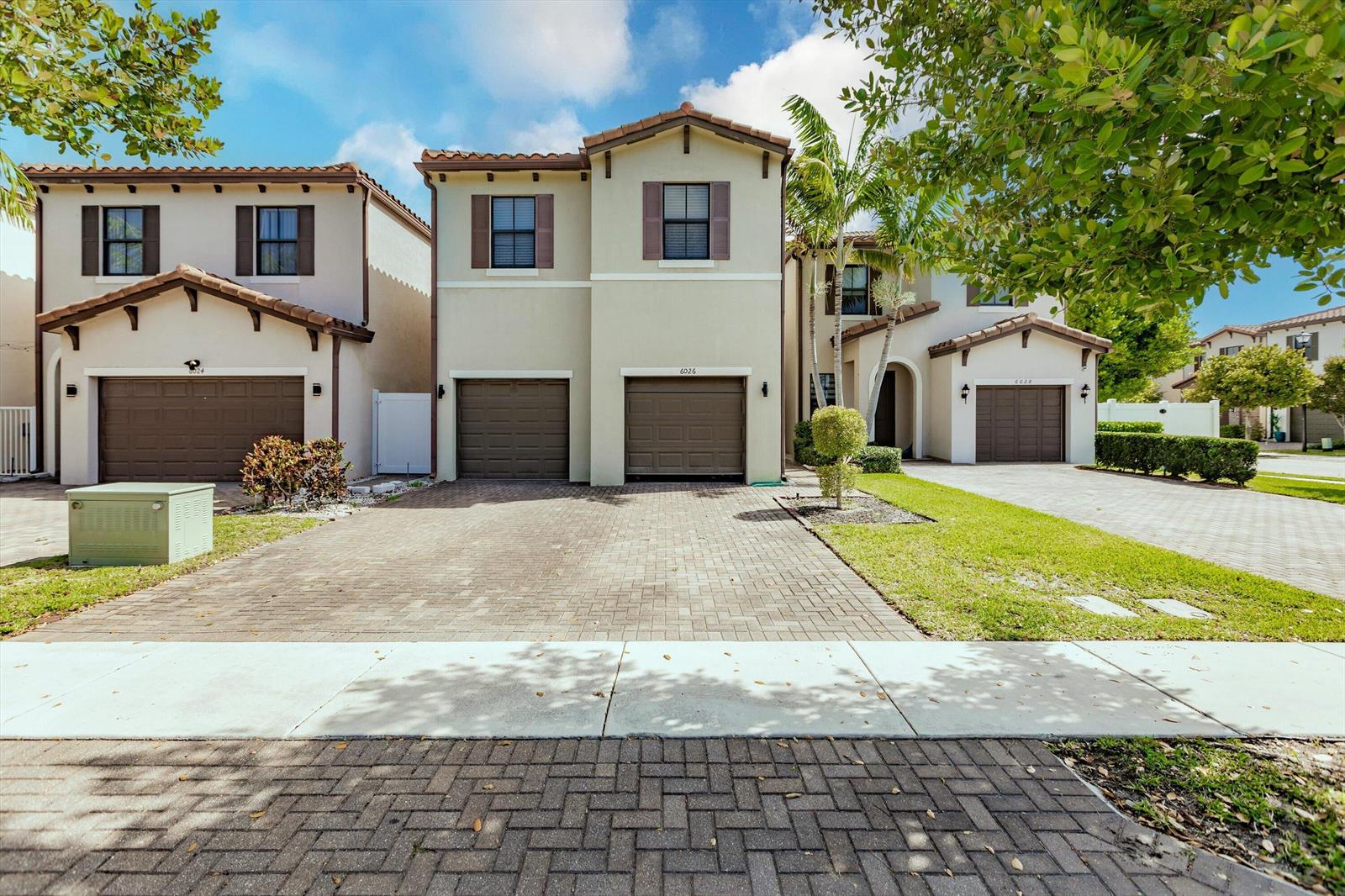 A street-level view of three attached two-story townhomes with paved driveways and brown garage doors — 6026 Pine Tree Way # 6026, Riviera Beach FL 33410, RIVIERA BEACH, FL