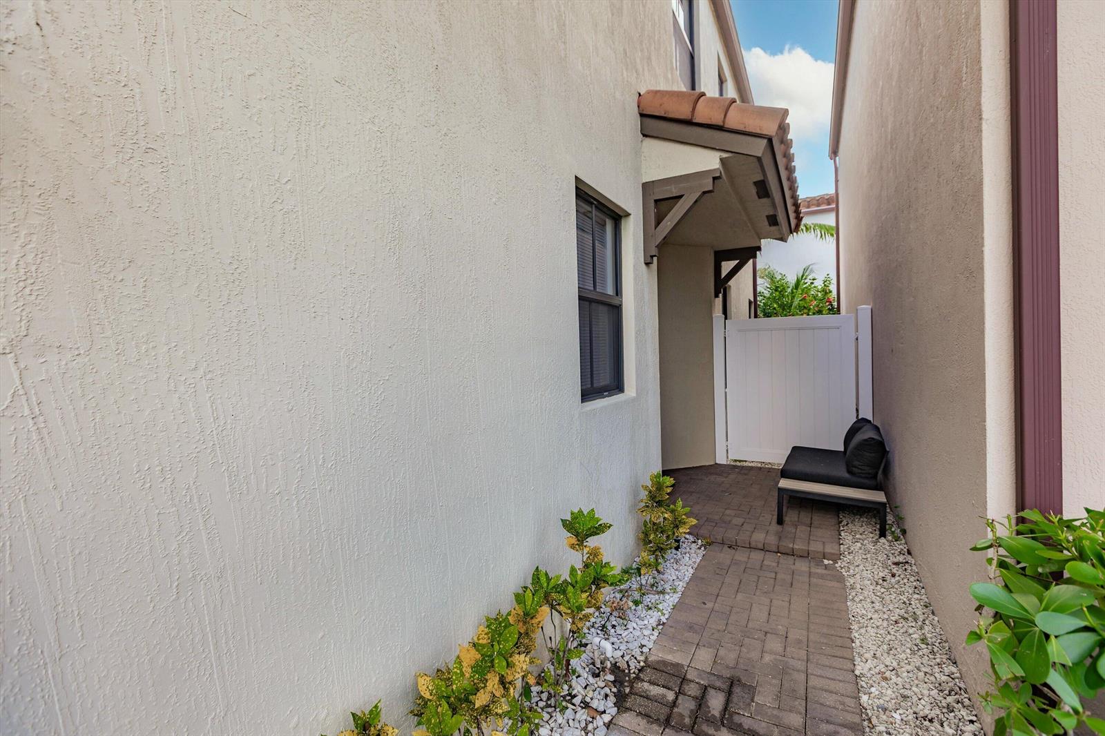 A narrow paved walkway leads between two stucco buildings toward a white gate and bench — 6026 Pine Tree Way # 6026, Riviera Beach FL 33410, RIVIERA BEACH, FL