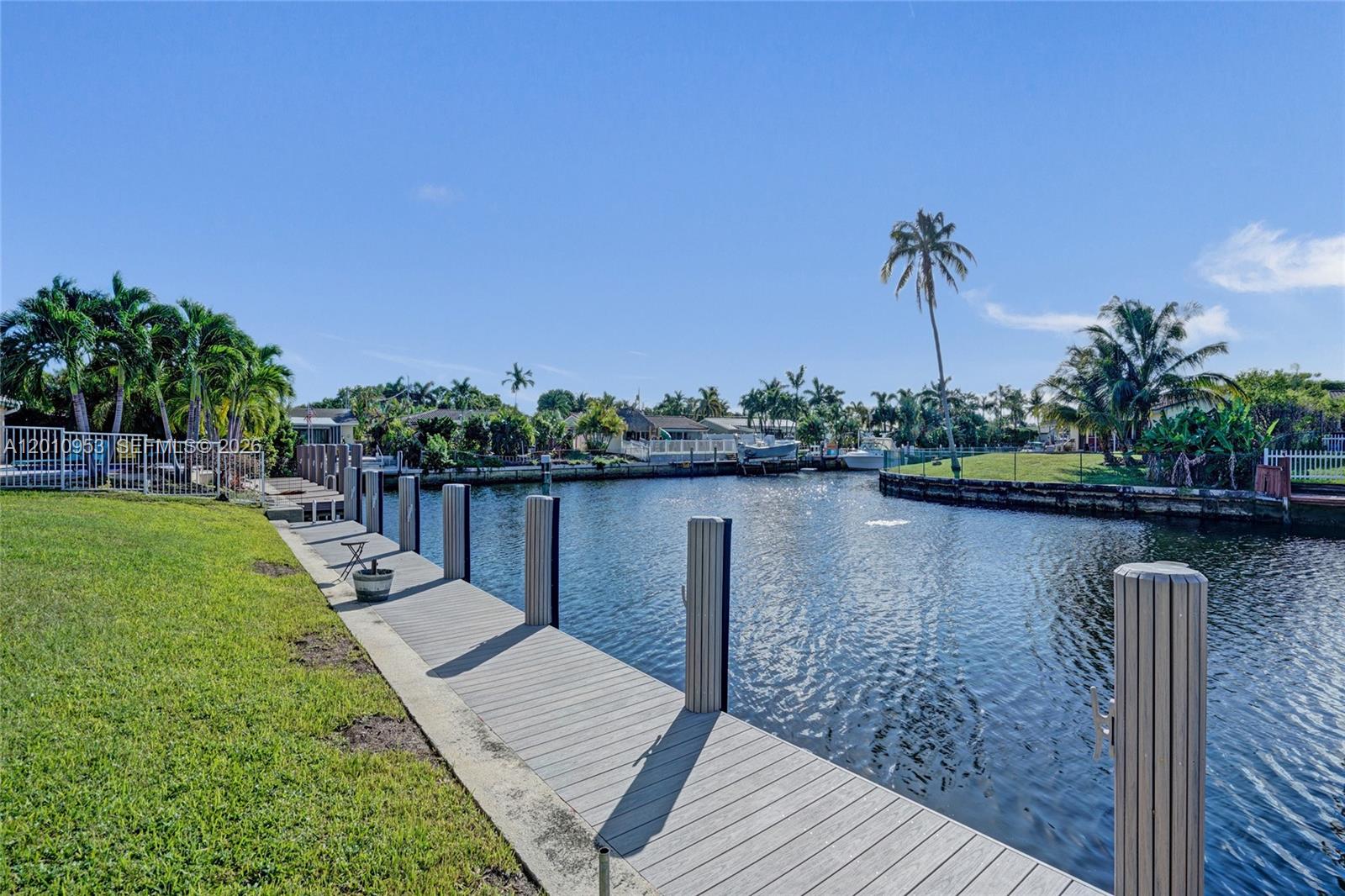 A wooden dock extends along a grassy bank beside a canal with palm trees — 949 SE 10th Ct, Pompano Beach FL 33060, POMPANO BEACH, FL