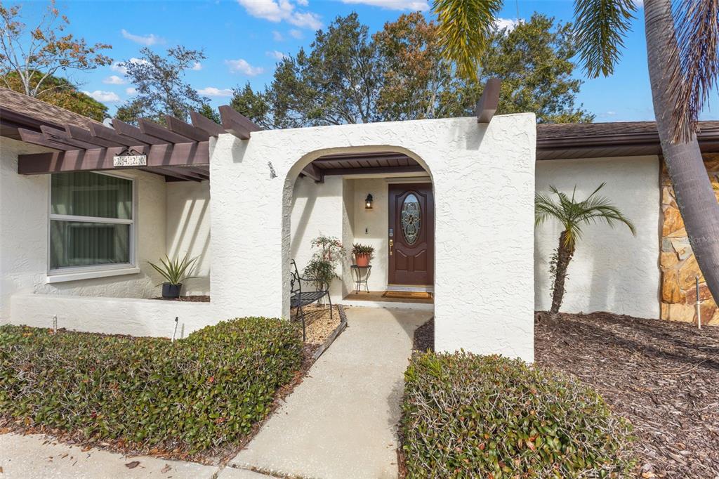The exterior of a house features a white stucco facade, a brown pergola, and an arched entryway — 3420 HYDE PARK DRIVE, CLEARWATER, FL