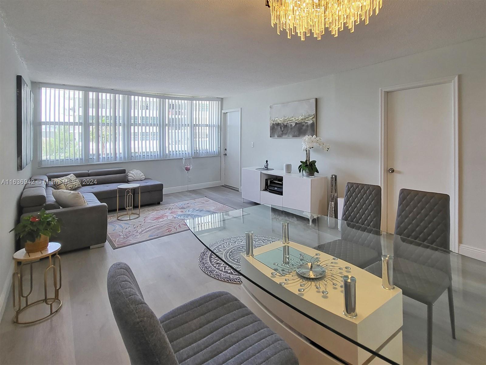 A living and dining area features a glass table, grey sofa, and large window blinds — 1825 S Ocean Dr # 206, Hallandale Beach FL 33009, HALLANDALE BEACH, FL