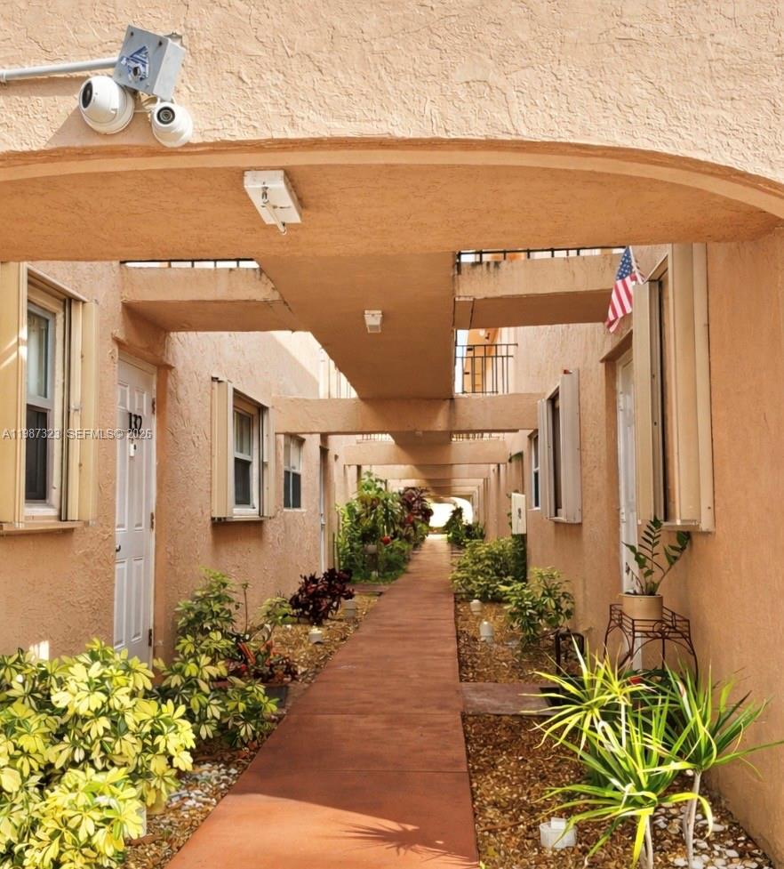 An outdoor walkway between two rows of apartment units with plants and a security camera — 550 SW 2nd Ave # 1210, Boca Raton FL 33432, BOCA RATON, FL