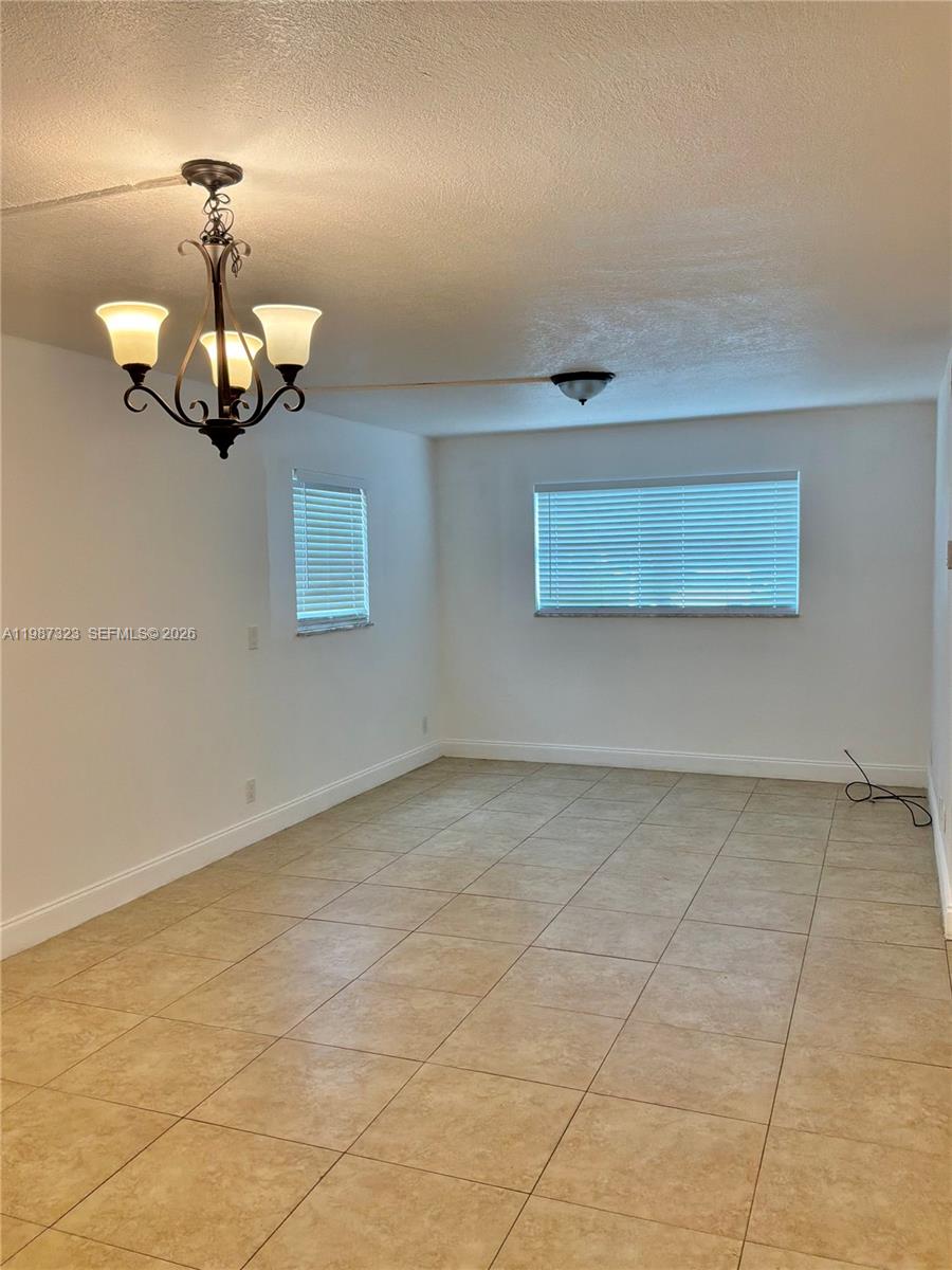 An empty room features white walls, beige tile flooring, a chandelier, and two windows — 550 SW 2nd Ave # 1210, Boca Raton FL 33432, BOCA RATON, FL
