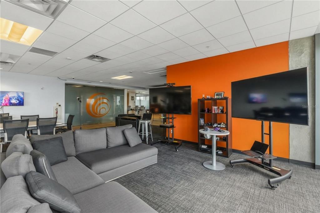 A modern office lounge features a gray sectional sofa, two large televisions on stands, and orange walls — 189 S ORANGE AVENUE UNIT 1950S, ORLANDO, FL