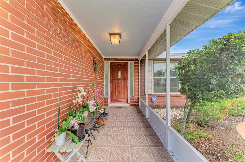 A covered brick-walled porch features a wood front door, tiled flooring, and potted orchids — 1460 CORTES DRIVE, ENGLEWOOD, FL