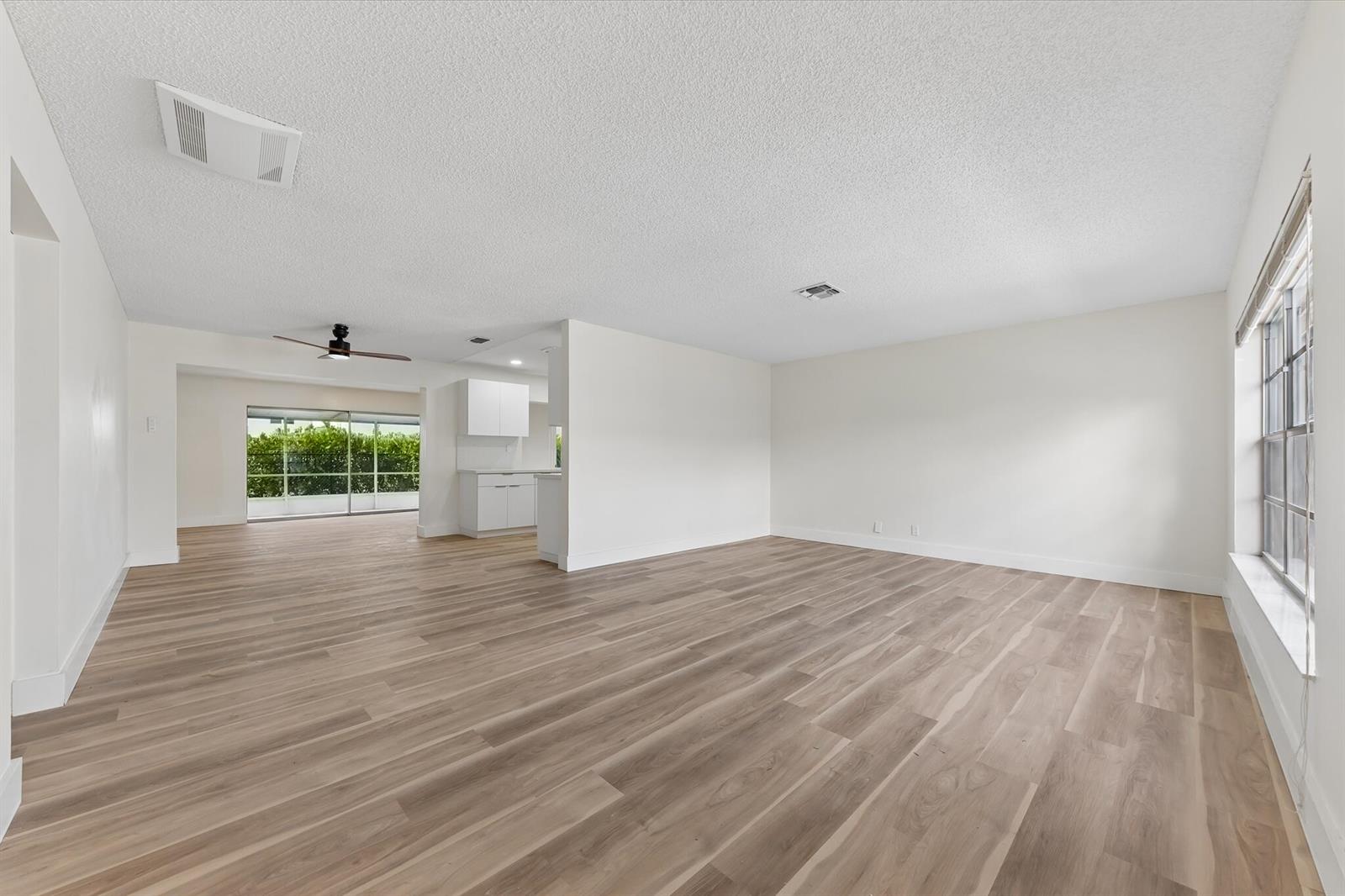 An empty living room with light wood-look flooring, white walls, and a view of a kitchen — 7198 E Pine Forest Cir Circle E, Lake Worth FL 33467, LAKE WORTH, FL