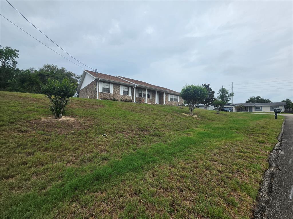 A single-story brick house sits on a sloped grassy lot under a cloudy sky — 1732 TOPAZ TERRACE, DELTONA, FL