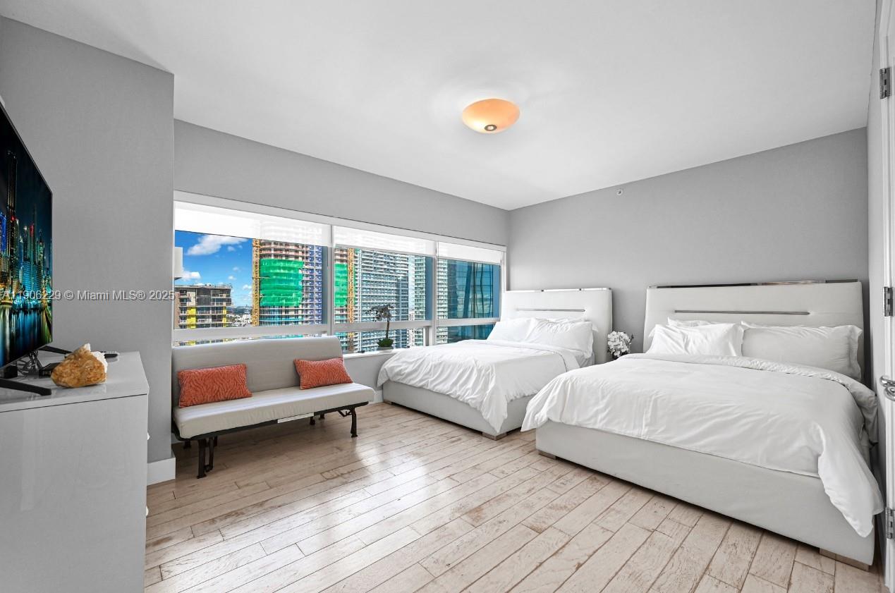 A bedroom features two white beds, a sofa, and large windows overlooking a city skyline — 1435 Brickell Ave # 3401, Miami FL 33131, MIAMI, FL