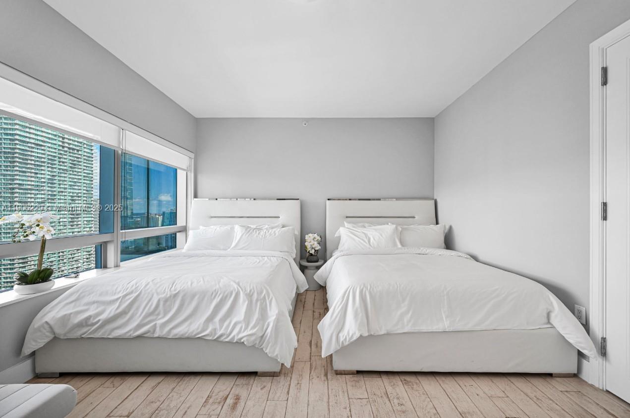 A bedroom features two white beds, light wood-look flooring, and floor-to-ceiling windows with city views — 1435 Brickell Ave # 3401, Miami FL 33131, MIAMI, FL