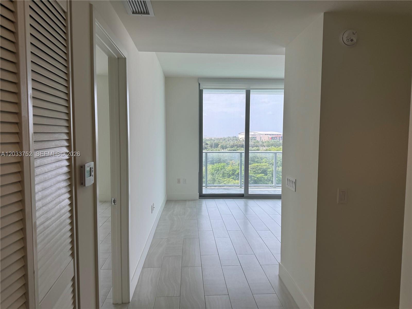 A bright room with light tile flooring leads to a balcony overlooking trees and buildings — 2000 Metropica Way # 810, Sunrise FL 33323, SUNRISE, FL