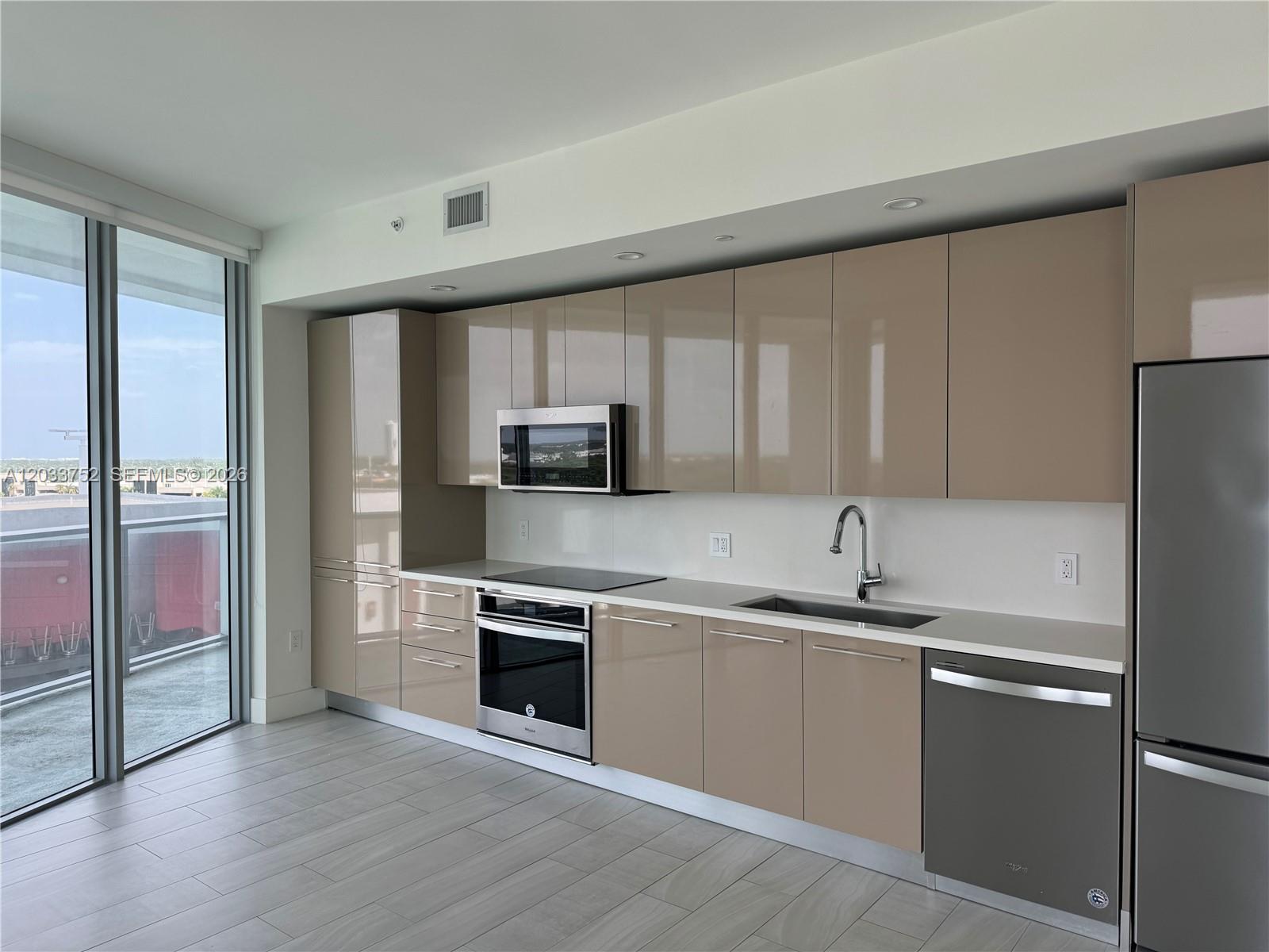 A modern kitchen features glossy beige cabinets, stainless steel appliances, and a balcony view — 2000 Metropica Way # 810, Sunrise FL 33323, SUNRISE, FL