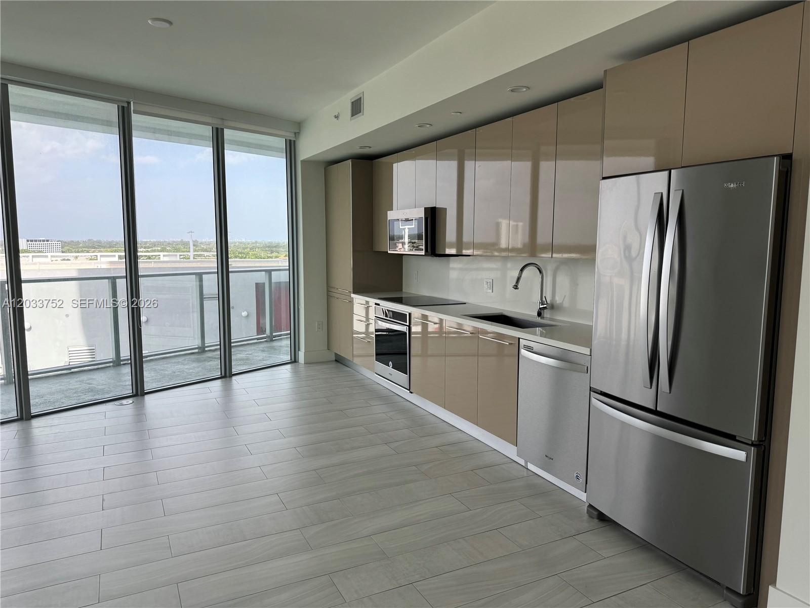A modern kitchen features glossy beige cabinetry, stainless steel appliances, and a balcony with views — 2000 Metropica Way # 810, Sunrise FL 33323, SUNRISE, FL