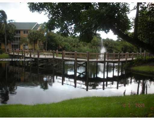 A wooden boardwalk crosses a pond with a fountain in front of a multi-story building — POMPANO BEACH, FL