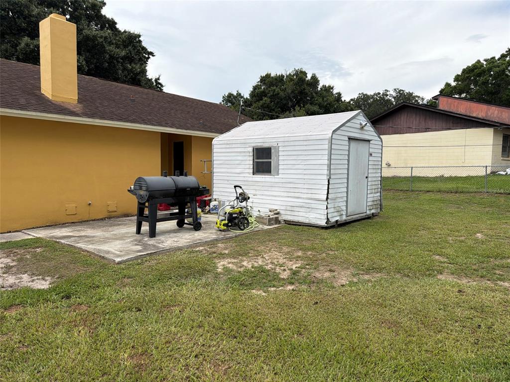 A backyard features a yellow house, a concrete patio, a grill, and a white shed — 7023 FOX CHASE DRIVE, LAKELAND, FL