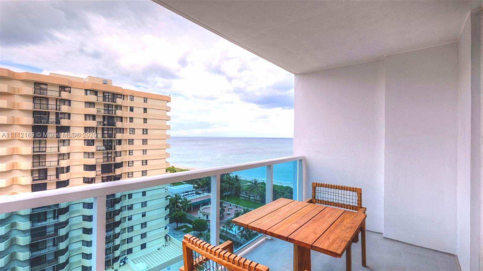 A balcony with a wooden table and chairs overlooks the ocean and a neighboring building — 102 24th St # 1211, Miami Beach FL 33139, MIAMI BEACH, FL