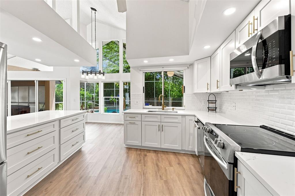 A modern white kitchen features quartz countertops, stainless steel appliances, and large windows overlooking a pool — 13513 WOODSIDE DRIVE, HUDSON, FL