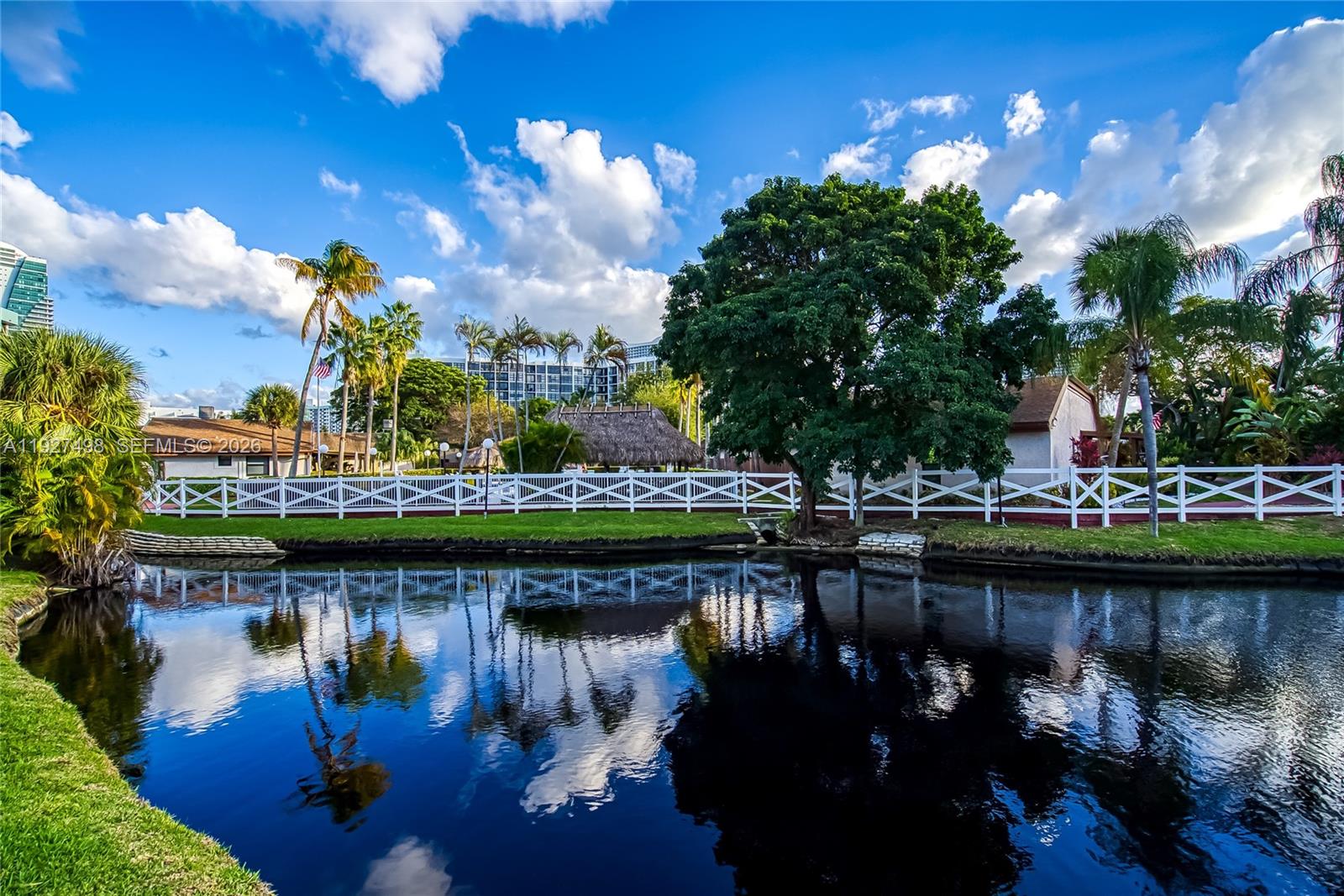 A white fence borders a pond with reflections of palm trees and a large tree — 2669 S Parkview Dr, Hallandale Beach FL 33009, HALLANDALE BEACH, FL