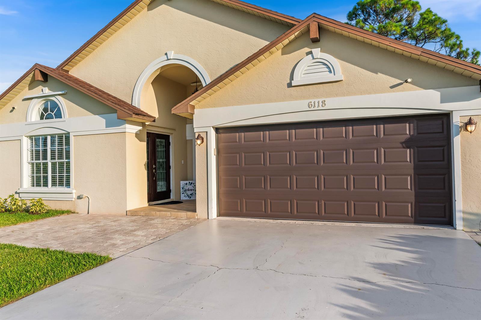 A tan house exterior features a brown garage door, arched entryway, and a paved driveway — 6118 Pompano Street, Jupiter FL 33458, JUPITER, FL