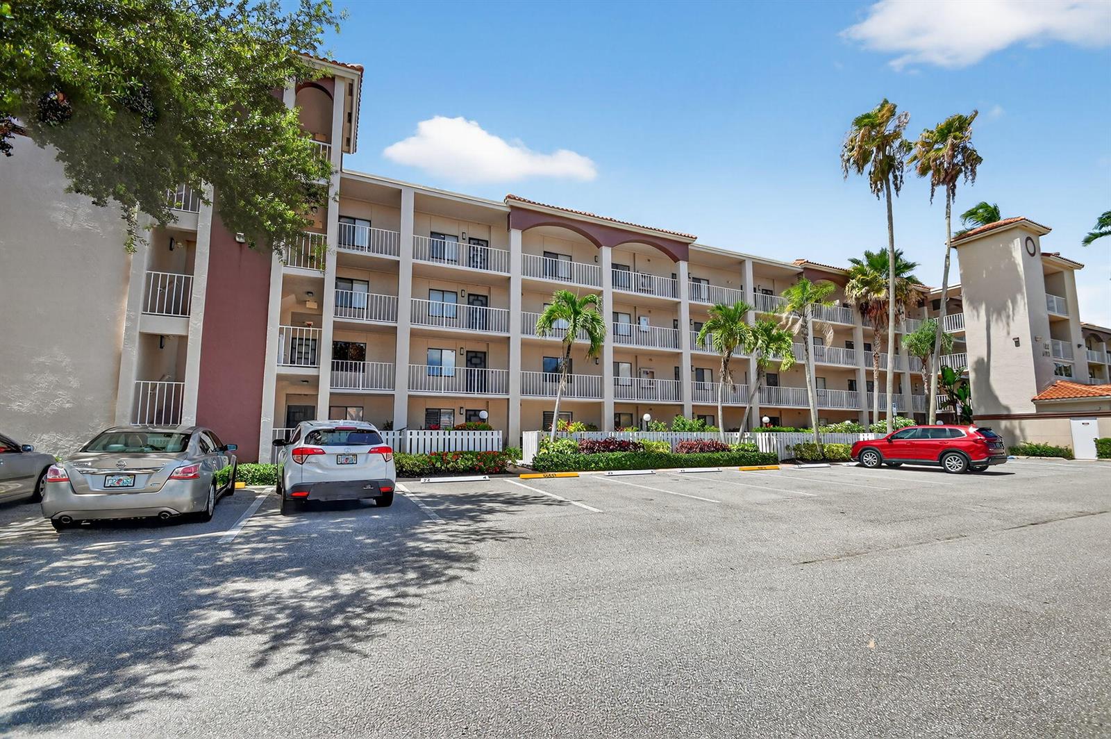 A multi-story apartment building with balconies stands behind a paved parking lot with several cars — 6193 Pointe Regal Circle # 402, Delray Beach FL 33484, DELRAY BEACH, FL