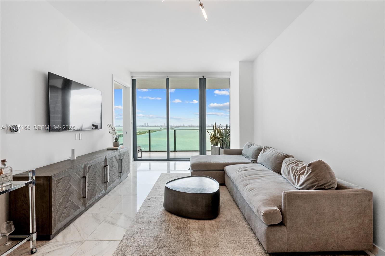 A modern living room features a grey sectional sofa, wooden media console, and balcony view — 480 NE 31st St # 2405, Miami FL 33137, MIAMI, FL