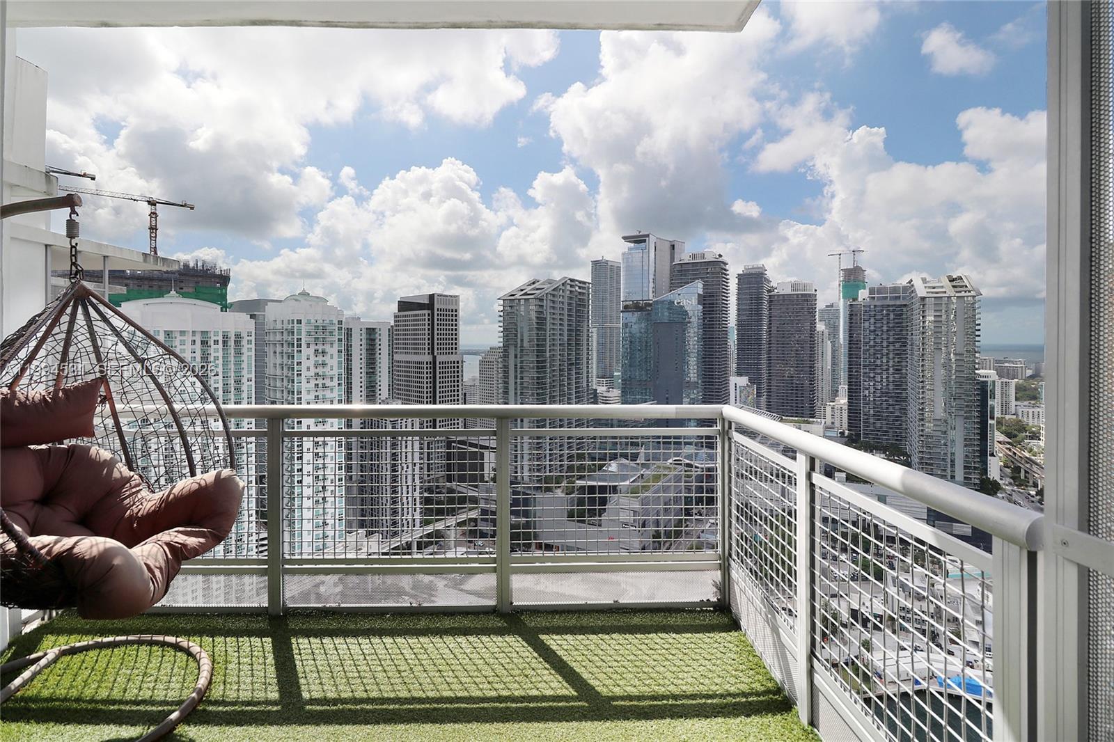 A high-rise balcony with artificial turf flooring features a hanging chair overlooking a city skyline — 350 S Miami Ave # 4110, Miami FL 33130, MIAMI, FL