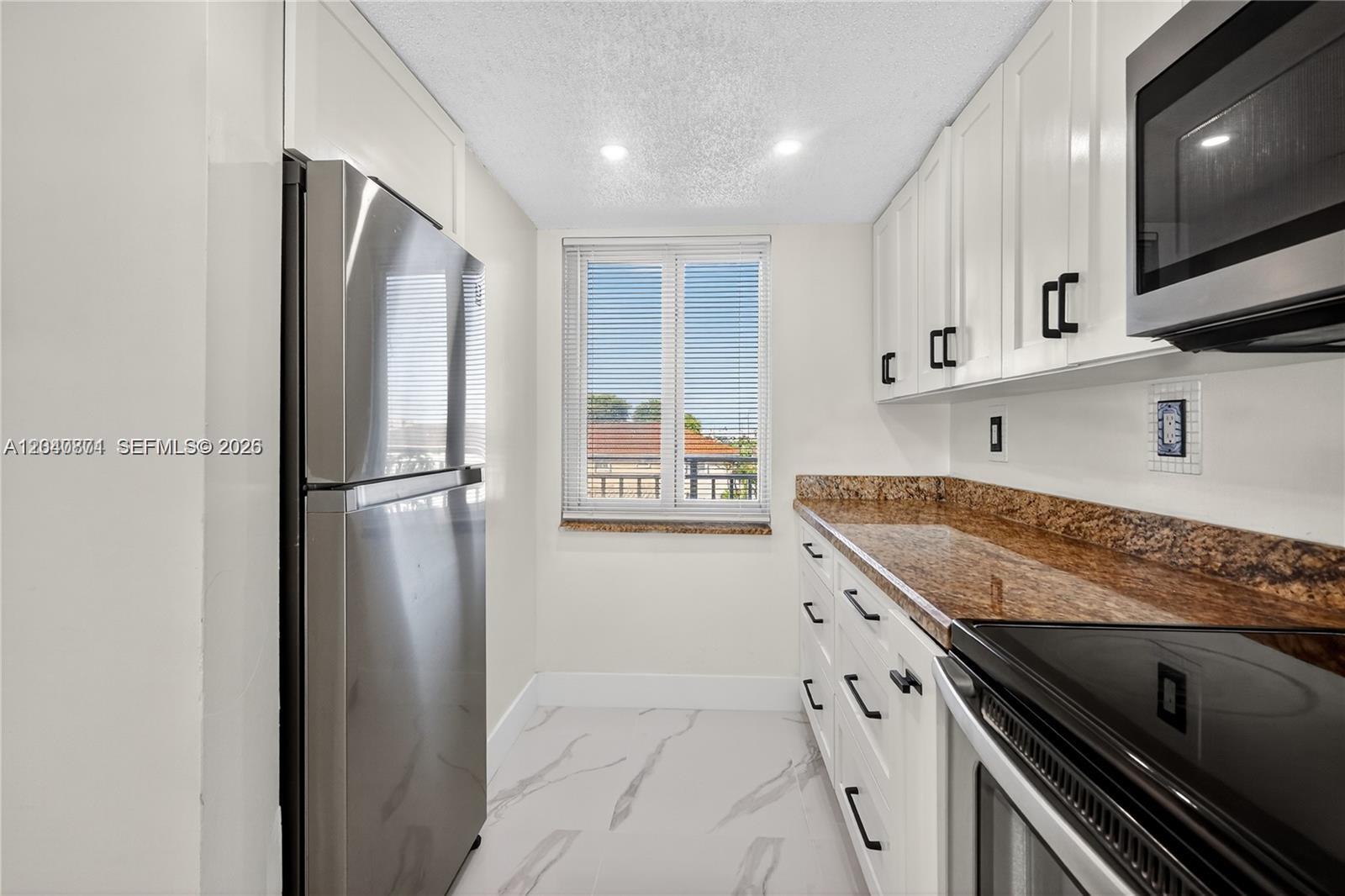 A galley kitchen features white cabinets, granite countertops, stainless steel appliances, and marble-look tile flooring — HIALEAH GARDENS, FL