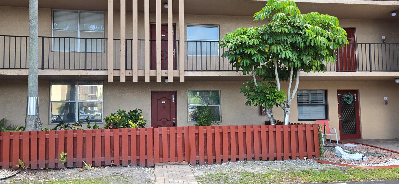 A two-story apartment building exterior features a red wooden fence and a large green tree — 7300 Lake Cir Drive # 107, Margate FL 33063, MARGATE, FL